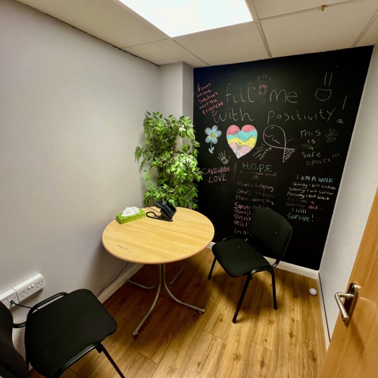 the photo shows an inside room from the Stevenage Crisis Cafe, including chairs and a table, a plant, plus a black wall with chalk artwork and writing