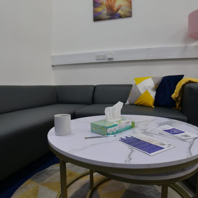 the photo shows an inside room from our Hatfield Crisis Cafe, which includes a long sofa and cushions, a table with leaflets and flyers, a lamp, art on the walls